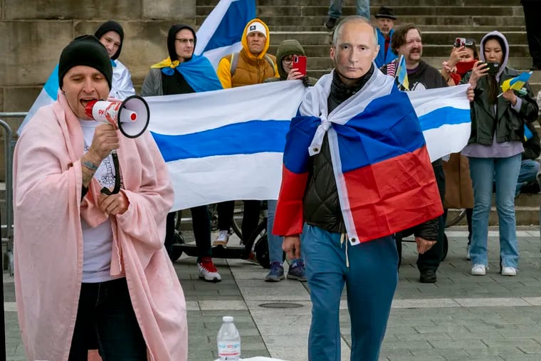 Alexander Kashapov (left), who is Russian, leads a chant “arrest Putin”  as a fellow demonstrator draped in a Russian flag wears a mask of the Russian leader Vladimir Putin. 