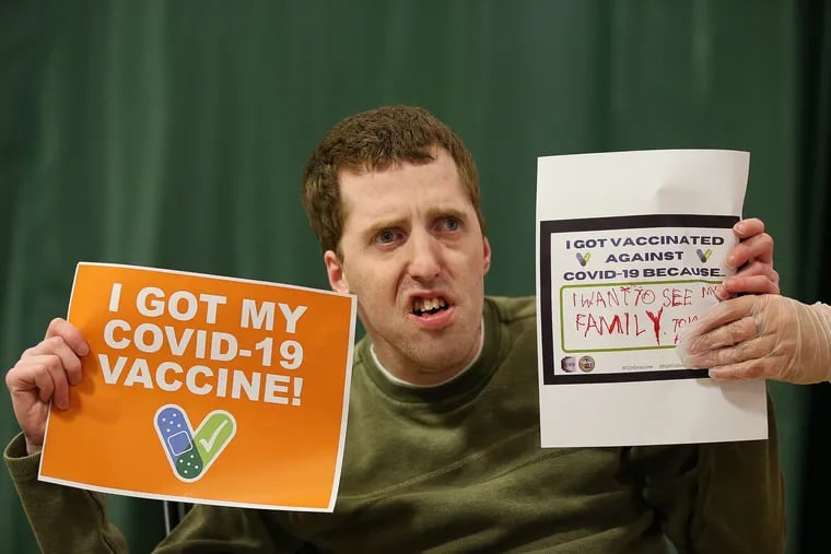 Resident Tom McMullan celebrates by holding up two signs after he received the COVID-19 vaccine at Melmark in Berwyn on Wednesday. The sign on the right gives the reason he wanted to receive the vaccine. It reads, “I want to see my family.”