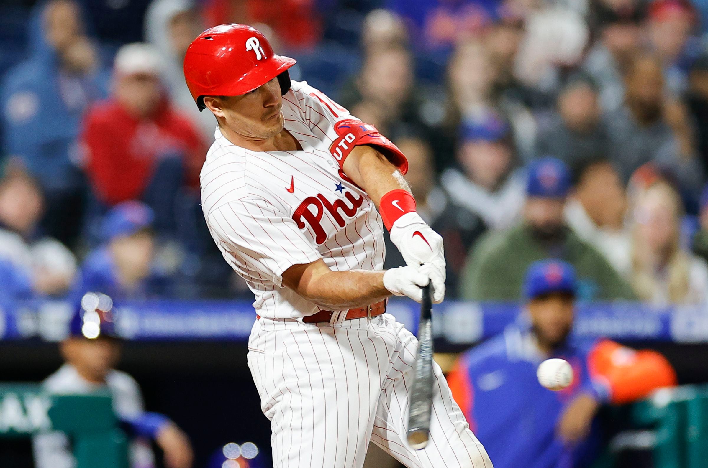 Philadelphia Phillies - Alec Bohm smiling, while putting on his white and  red batting gloves. He is holding a baseball bat underneath his left arm.  Bohm is wearing a red long sleeve