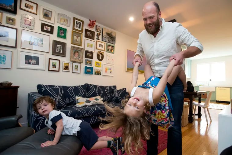 Taylor Foxwell plays with two of his children, Wally and Lady, in the living room of their Fishtown home. Decor includes a gallery wall of family photos, quirky figurines, and children's art.